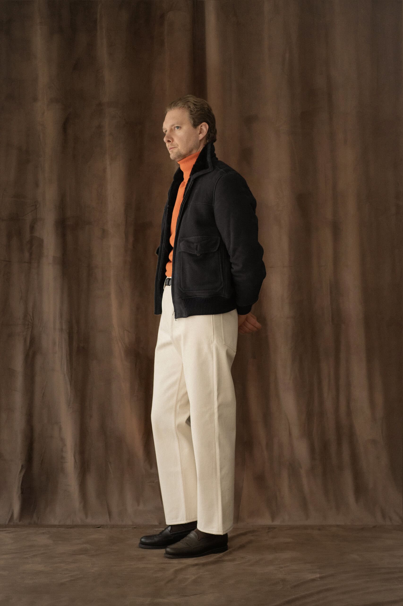 Dunkle Fliegerjacke mit orangefarbenem Rollkragenpullover und cremefarbener Hose